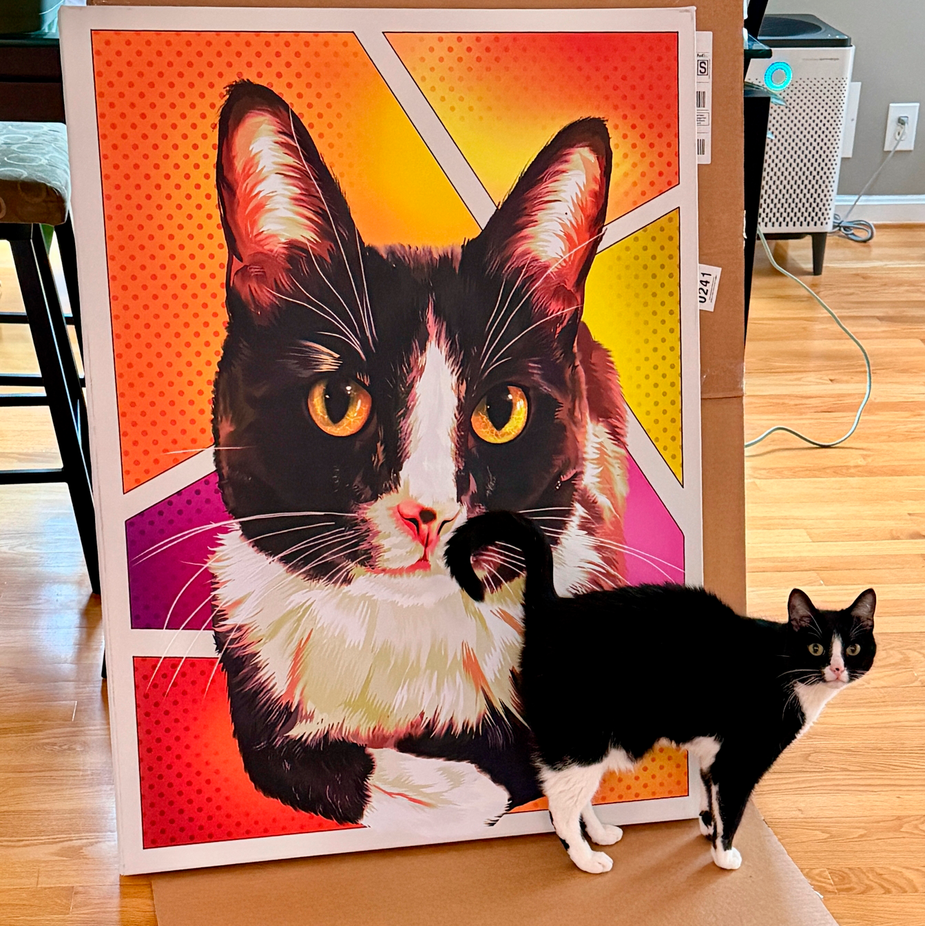 Black and white cat standing next to a large framed picture of a similar cat on a wooden floor.